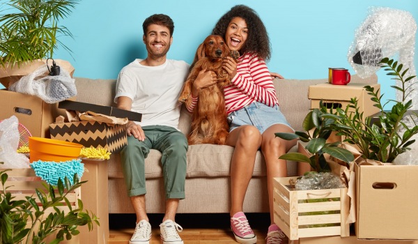 A couple sitting on their couch with their dog, surrounded by moving boxes
